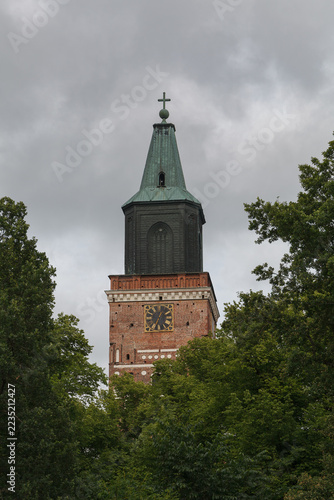 Turku Cathedral spire rises between leafy branches under an overcast summer sky.