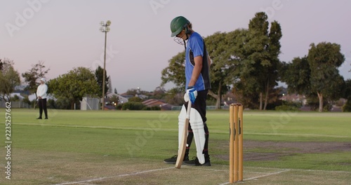 Teenage batsman adjusting bat grip at cricket ground as bowler approaching then walking off pitch