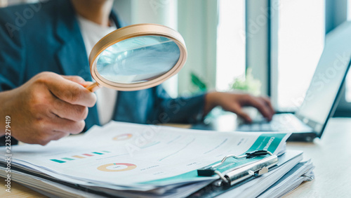 Close view of a magnifying glass over business reports as an office worker takes notes beside a laptop, illustrating document review, financial analysis, verification, and detailed auditing processes.