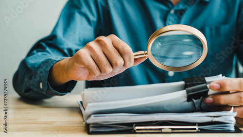 Office worker examines a stack of documents with a magnifying glass at a desk. Close view of hands and paperwork illustrates auditing, compliance review, record verification, and attention to detail.