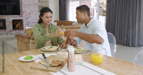 Diverse couple in button-up shirts sitting at wooden dining table, clinking orange juice glasses