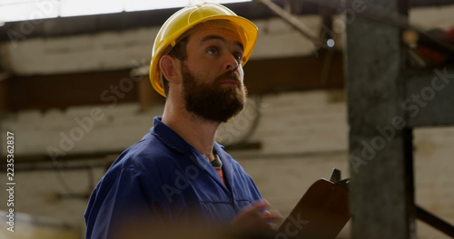 A focused man in workwear and hard hat with clipboard likely evaluates a construction site.