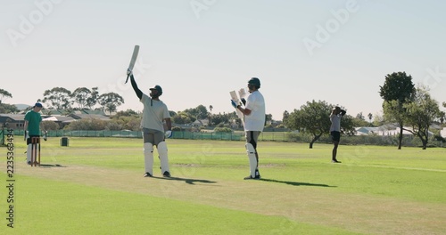 Male batsman is raising bat on suburban park pitch while wearing helmet near wicket stumps