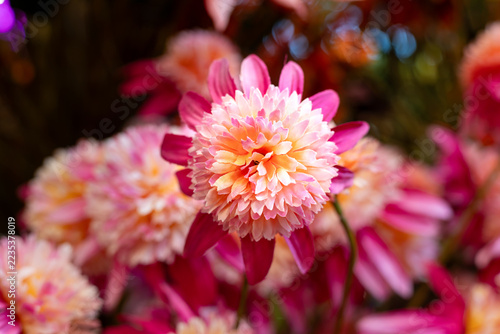 Close-up of an artificial chrysanthemum-style flower with layered pink, peach and cream fabric petals, surrounded by softly blurred colorful blooms in a delicate floral arrangement.