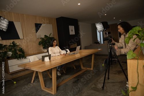 Indian host at wooden table in studio presenting on laptop, mic while African tech operating DSLR