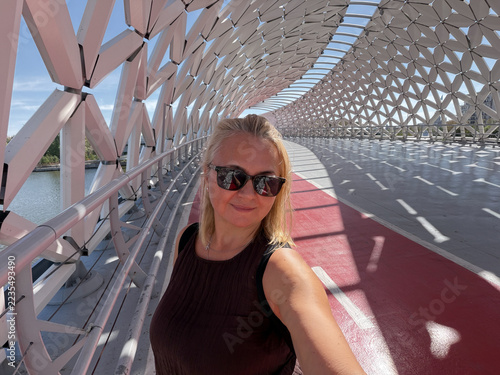 Atyrau Bridge. Futuristic pedestrian bridge in Astana, woman tourist captures travel experience beneath geometric steel canopy with red walking path and modern architecture, Kazakhstan