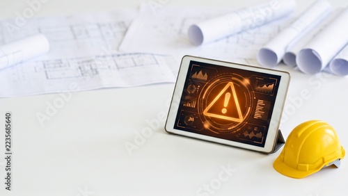 Tablet showing warning triangle beside blueprints and yellow hard hat for construction risk alerts, site safety monitoring and engineering project planning