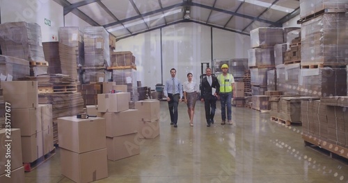 Four adults walking through warehouse aisle wearing hard hats beside stacked boxes on pallets