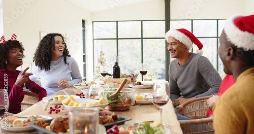 Diverse friends celebrating, sharing roast and wine at dining table, wearing Santa hats