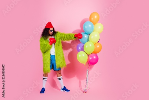 Fotografie Man in green fur coat and red boxing gloves punches colorful balloons on a pink