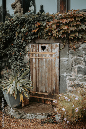 Petite porte en bois avec ouverture en forme de cœur dans un mur en pierre recouvert de lierre