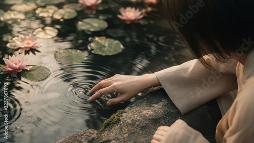 Woman touching water surface creating ripples near pink lotus flowers in pond